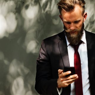 A bearded man in a red tie is intently looking at his cell phone, engaged in a conversation or checking messages.